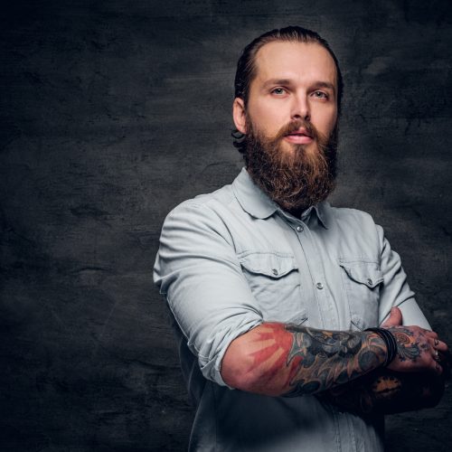Groomed bearded man with tattooes is posing at dark photo studio.