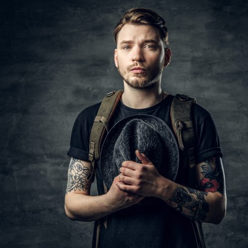 Portrait of a handsome tattooed traveler hipster male holds tweed hat in his arms and backpack on his back.
