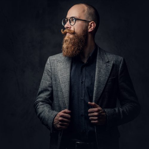 Portrait of redhead bearded male in eyeglasses dressed in an elegant wool suit over grey background.